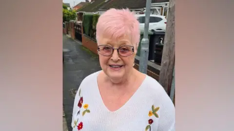 Jade Fenton A woman stands in a road outside a row of houses. She is wearing a white jumper which has flowers on and has short hair and is wearing glasses. 