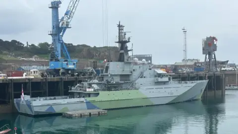 A large military ship painted grey, green, and blue is in a marina. Cranes and other harbour equipment fill the background.