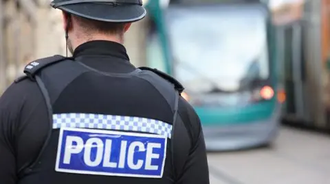 A police officer stands with his back to the camera. He is wearing a black uniform which includes a vest with the word "police" written on it. A Nottingham tram can be seen in the background.