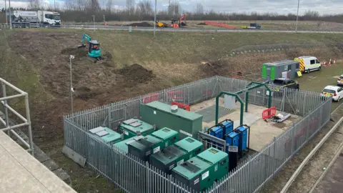 Tony Fisher/BBC A pumping station by a main road with a digger working on the embankment behind it. 