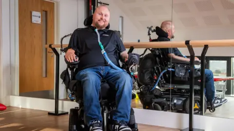 Matt Hampson Foundation Matt Hampson is sat in his wheelchair in one of the rehabilitation halls at The Matt Hampson Foundation. He is smiling. He is wearing a black shirt and blue jeans. He also has a ventilator in his neck which he breathes through. Behind him is a wooden bar used to help people in the hall, and there is a wooden floor. 
