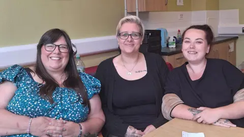 Karen (left) wears a bright blue flowery top and dark glasses. She has long dark hair. Louise has short blonde hair and wears a black top and cardigan, as well as dark framed glasses. Shannon also wears a dark top and has dark hair scraped back in a bun ponytail.  
