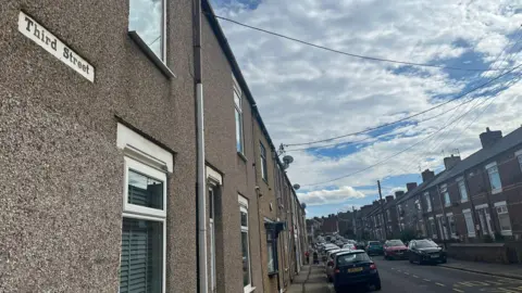 A view of a street of terraced houses with rows of cars parked on both sides of the street. A sign attached to the wall of the nearest house reads "Third Street".