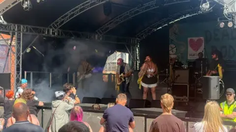 A woman can be seen on stage in a white and black short dress with a flowy top over it. She is singing into a microphone with other musicians including guitarists and a drummer alongside her. People can be seen standing at the front of the stage watching.