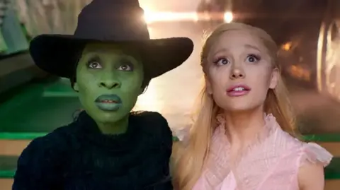 Universal Cynthia Erivo is pictured with a green face and witch's hat, next to Ariana Grande, who has a pink dress on and blond hair