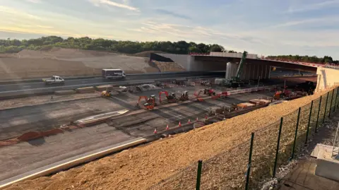 A shot of the A417 Missing Link project in Gloucestershire. It is a newly built road and traffic cones and diggers can be seen on the roadside. There is a bridge over the road.