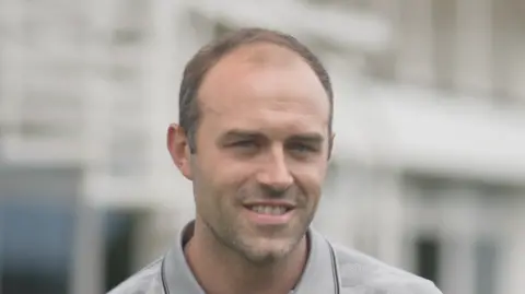 Jamie Niblock/BBC Jonny Sharkey smiles at the camera. He has thin dark hair that is receding. He wears a grey polo top.