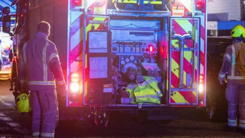 A night-time image of a Kent Fire & Rescue fire engine from the rear with a couple of fire officers facing away from character in uniform