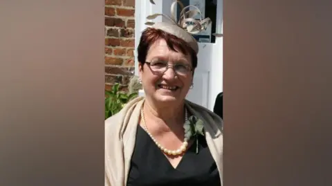 A woman is smiling while standing in front of the door of a house. She is dressed in formal wear, with a black top or dress, cream-coloured pashmina, a white or cream-coloured rose buttonhole, pearl necklace and a cream coloured pillbox hat with fascinator detail. She is also wearing glasses and she has red-brown short hair.
