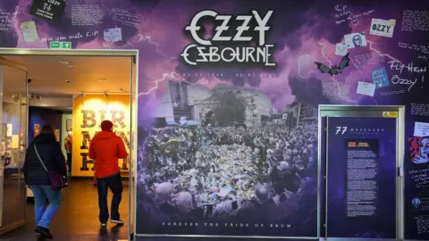 PA Media Two people walk past a museum stand for the Ozzy Osbourne exhibition, which depicts his funeral parade and tributes. A female visitor is wearing a black coat and jeans and the male a red coat, red hat and jeans.