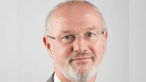 Gloucestershire County Council Headshot of Stephen Davies wearing a grey suit with a white shirt and multi-coloured tie