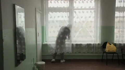 Pavel Talankin/BBC Storyville/Made in Copenhagen Pasha behind a net curtain sticking an X symbol on the window. chair with yellow bag. On left hand side a mirror. Green walls