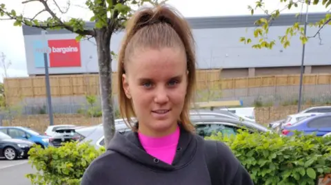 Family handout Holly Newton, photographed in a car park with trees and vehicles behind her, has long dark hair tied up in a pony tail and is wearing a pink shirt under grey hoodie. She is looking directly at the camera