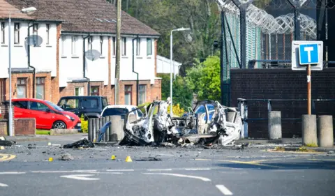 Pacemaker The aftermath of an explosion outside a police station. The wreckage of a car is shown, it has sustained significant fire damage. There is a police station in the background and a row of houses on the opposite side of the street. Debris is strewn across the entrance to the street