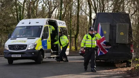 A police van parked on a road surrounded by trees with its side open. There are uniformed officers in and around the van, and another person walking around the back of a separate black van, which has a union flag attached to the back of it.