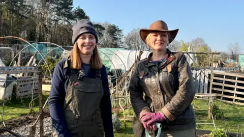 Two smiling women stood in an allotment holding gardening equipment 