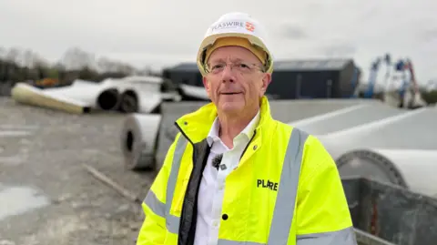 Andrew Billingsley, a man wearing frameless glasses, a white helmet, a yellow-high viz jacket and a white shirt, stands in an industrial yard looking at the camera. There is a collection of large wind turbine blades behind him. 