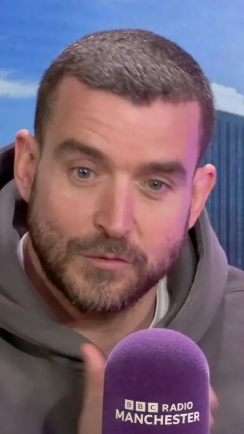 A man with short brown hair is sat behind a microphone which says BBC Radio Manchester