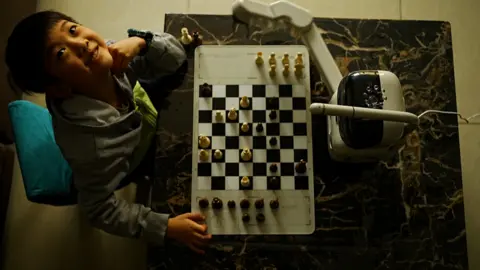 BBC/ Xiqing Wang looks on a smiling Timmy on camera. On the table, it has a chess board and a small robot with a white body and a black screen.     