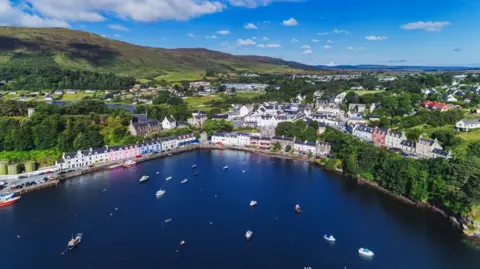 Getty Images An aerial view over a bay filled with small boats to Portree on Skye. The town has houses and other buildings among trees. Some of the houses have colourful exterior walls, including pink and blue. A green hill rises behind the house. It is a sunny day with white clouds in a blue sky.