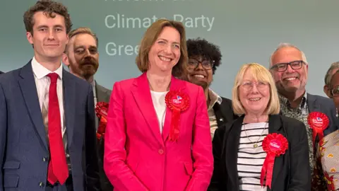 Ian Wyatt/BBC Six people standing on a stage and smiling. In the centre is Colchester MP Pam Cox, who is wearing a pink suit and a red rosette. She is standing with members of Colchester's Labour Party.