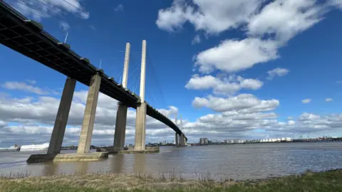 A wide low-angle shot of the Queen Elizabeth II Bridge (Dartford Crossing) stretching across the River Thames, under a bright blue sky with scattered white clouds.