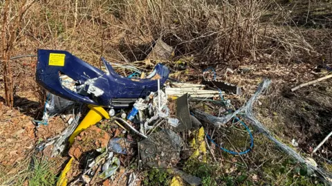 LDRS A blue car bumper and yellow plastic cabling is among a pile of rubbish on the ground, with grass around it.