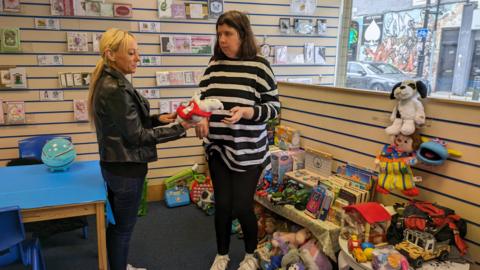 Two adult woman looking at a stuffed toy in a room filled with toys and books.