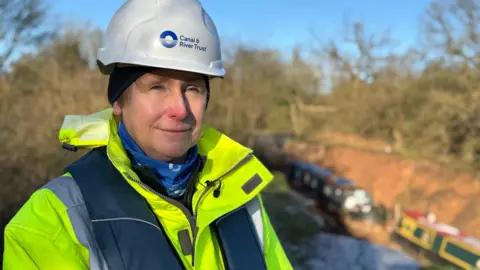 A woman in a white hard-hat which says Canal & River Trust on it. She wears a hi-viz jacket and stands in front of the site of the canal embankment collapse in Shropshire. Two boats can be seen in the hole left by the incident.