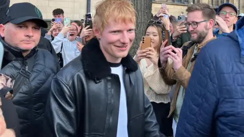 A ginger-haired man pushes through a crowd of people holding up phones. He is wearing a black leather jacket with fur around the collar and a white t-shirt.
