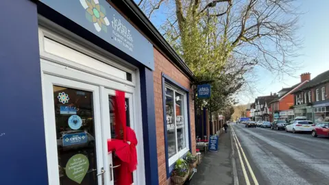 ELLEN KNIGHT/BBC An exterior photograph of the stop - which is a single-storey red brick building with dark blue window frames and a bright red ribbon tied around the shop door. Above the door, in light yellow letters, reads 'John R Thomas Florist', with the shop logo - a green flower - next to it. On the right of the photo, the Church Stretton high street stretches off into the distance. The sky is blue and there's bright sunshine. 