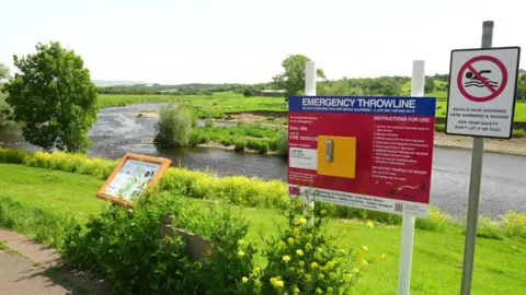 View from the banks of the River Ribble in Ribchester with signs detailing instructions in an emergency and a no swimming sign with a warning saying people have drowned in the river.