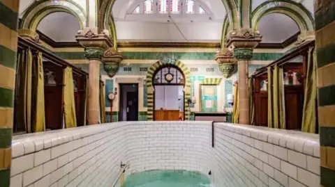 A highly decorated room with green and mustard tiles and ornate columns. There is a deep plunge pool in the foreground with white tiles. At the sides there are booths separated by wooden panels with decorations at the top and green privacy curtains.