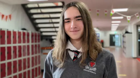 Alexia smiles into the camera. She is wearing a grey school jumper and has long brown hair. She is standing in a school hallway, with red lockers, stairs and Welsh flag bunting in the background. 