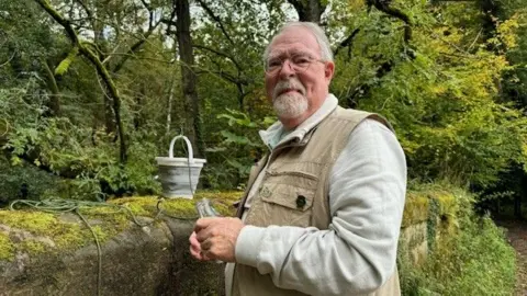 A man with a grey beard wearing a beige jacket looks towards the camera. He is standing next to a wall, behind the wall there are trees