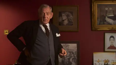 Actor Sir Ian McKellen is dressed in a black jacket with a white handkerchief in its breast pocket. He is also wearing a white shirt, grey cardigan and black tie. Lowry's paintings are on the scarlet-painted wall behind him.