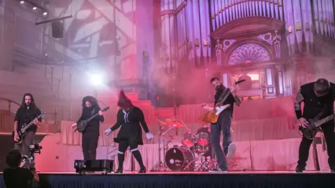 Five members of a rock band all dressed in black playing instruments in front of the smoke covered pipes of a church organ.