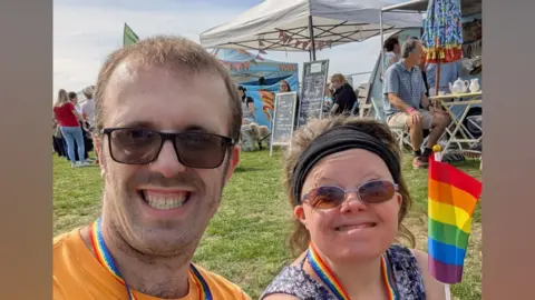 Halow Two people sit on grass at an outdoor event wearing lanyards and holding small rainbow flags, with food stalls, seating and other visitors in the background.