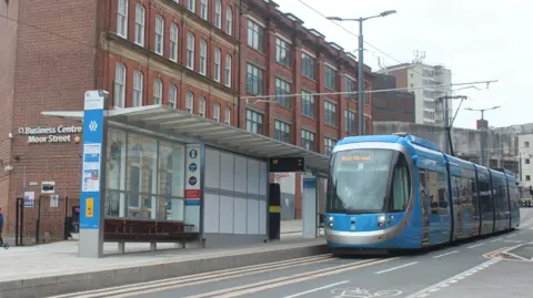 A blue and silver tram pulling up to a tram stop on a city street by a four-storey building with white lettering on the wall saying Business Centre Moor Street.