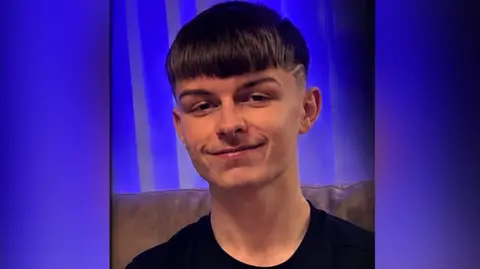 A teenage boy smiling at the camera. He has short brown hair and a black t-shirt on. 