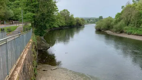 A winding stretch of the River Nith with gravelly banks and trees down either side with hills in the distance and hazy, cloudy, light blue sky overhead. There is a footpath on the left hand side of the river, and steps can be seen going down into the water. 