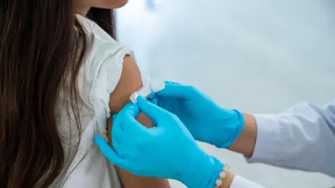 A young girl is has a plaster put on her arm by a person wearing blue gloves