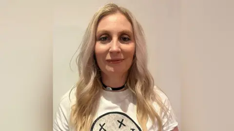 KidsAid A smiling Carla Managan looks directly at the camera as she is photographed standing in front of a white wall. She has long blonde hair and is wearing a white T-shirt with a design on it. She is wearing a black necklace. 