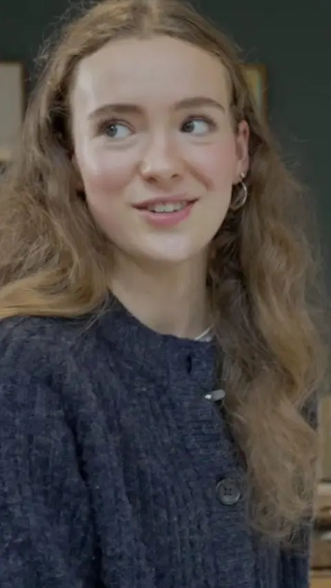 Lucy Sturt looks lsightly away from the camera. She has light brown long curly hair and wears hoop earrings and a navy blue marl knitted cardigan.