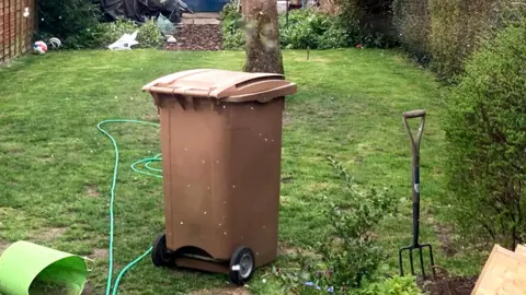 Matt Marvel/BBC A brown garden waste bin sits in a garden. A green hose pipe lies on the grass around it as well as a garden fork.