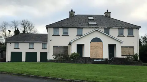 Clonduff parochial house which is a large residential building attached to parish offices. The ground floor windows of the vacant building have been boarded up.