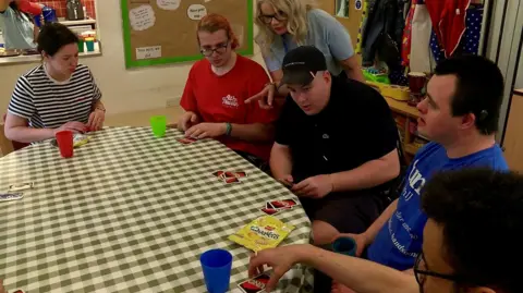 Dave Edwards/BBC A group of young people sit around a table playing cards