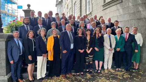 Members of the States of Guernsey. All 40 of them, a mix of men and women in business attire, are standing on the steps of the Royal Court. 