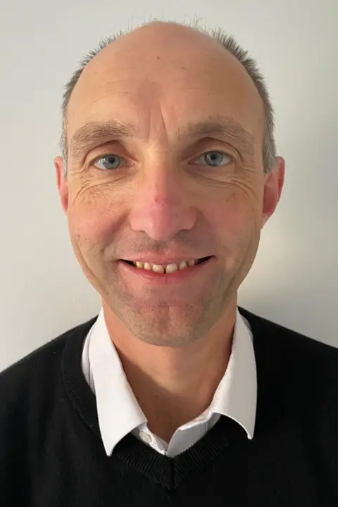 Dr Matthew Dominey Dr Matthew Dominey is smiling at the camera. He has short grey hair and blue eyes. He is wearing a white shirt under a black V-neck jumper.