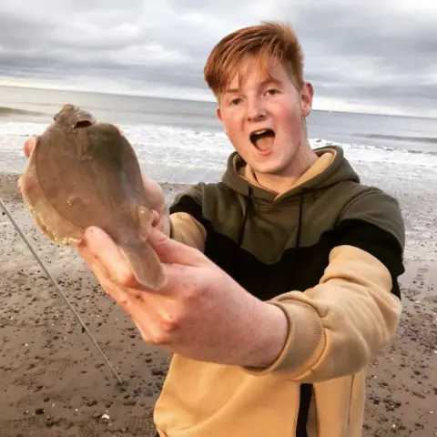 Karl Shannon A male teenager looks surprised as he holds up a dab - a brown flat fish - caught while angling. He is tall, has a mop of ginger hair and wears a beige, black and green hooded top. He is standing on a stony beach with the grey sea behind him. 